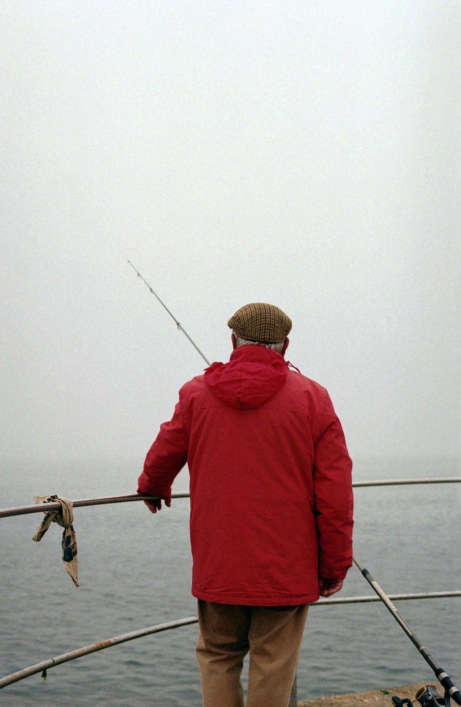 A man in a red jacket and plaid cap stands with a fishing rod overlooking the water on a hazy day.