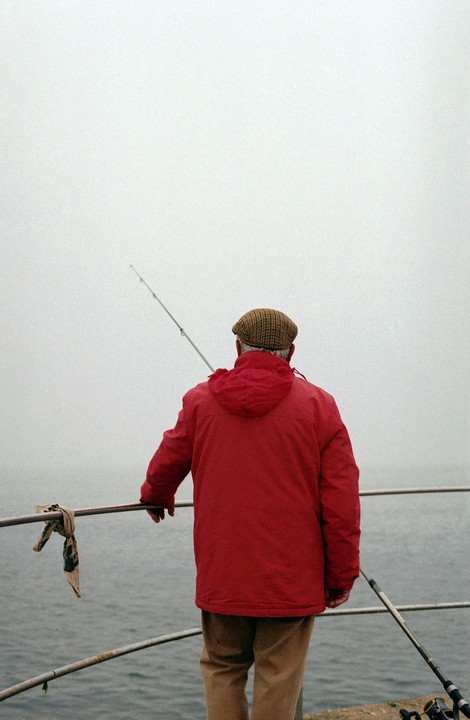 A man in a red jacket and plaid cap stands with a fishing rod overlooking the water on a hazy day.