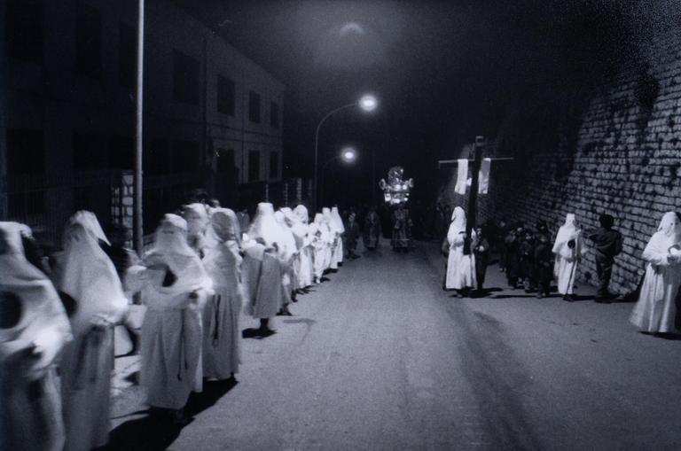A procession of people in white robes and hoods walk down a street at night, with some carrying a large cross and another a decorated float.