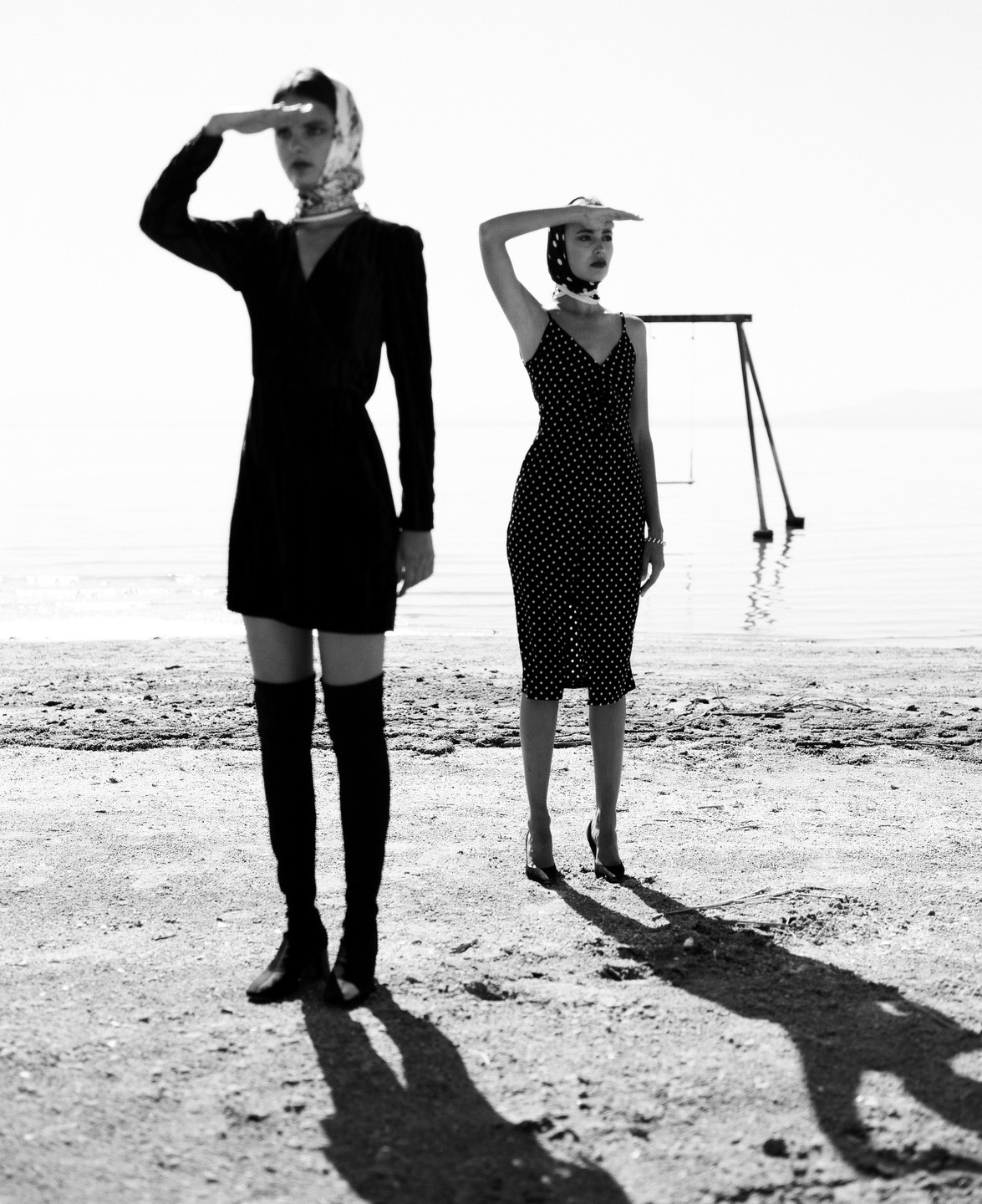 Two women stand on a beach with their hands shading their eyes from the bright light.