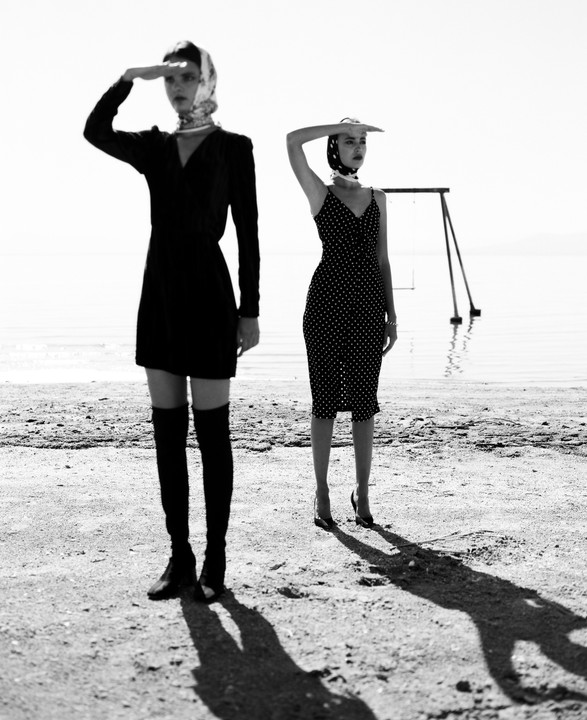 Two women stand on a beach with their hands shading their eyes from the bright light.