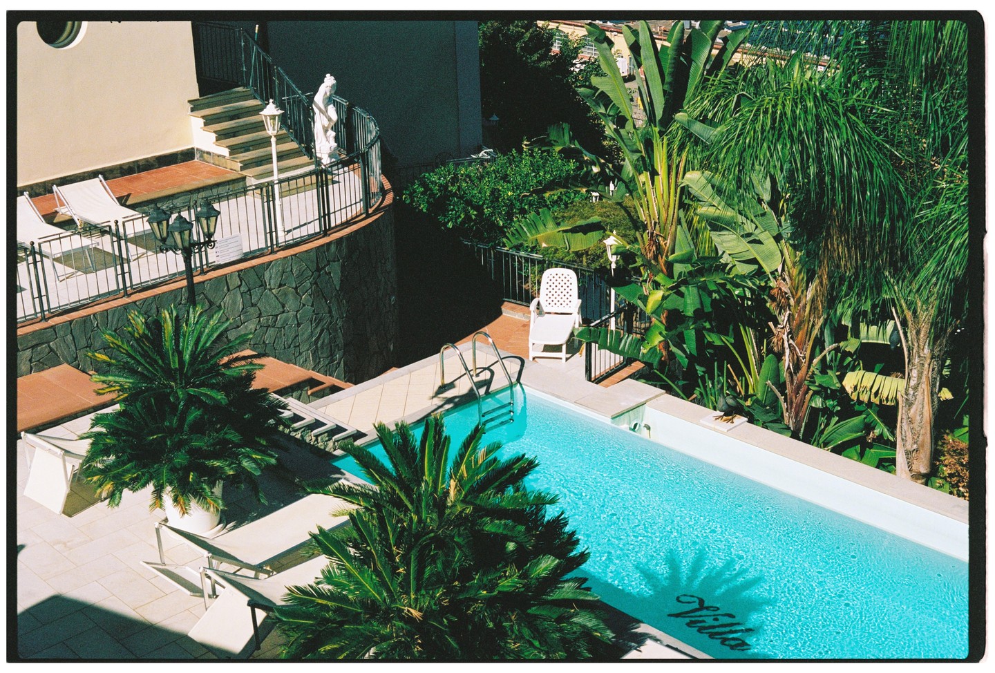 An elevated view shows a swimming pool with "Villa" written on its bottom, surrounded by palm trees and a building with a balcony and stairs.