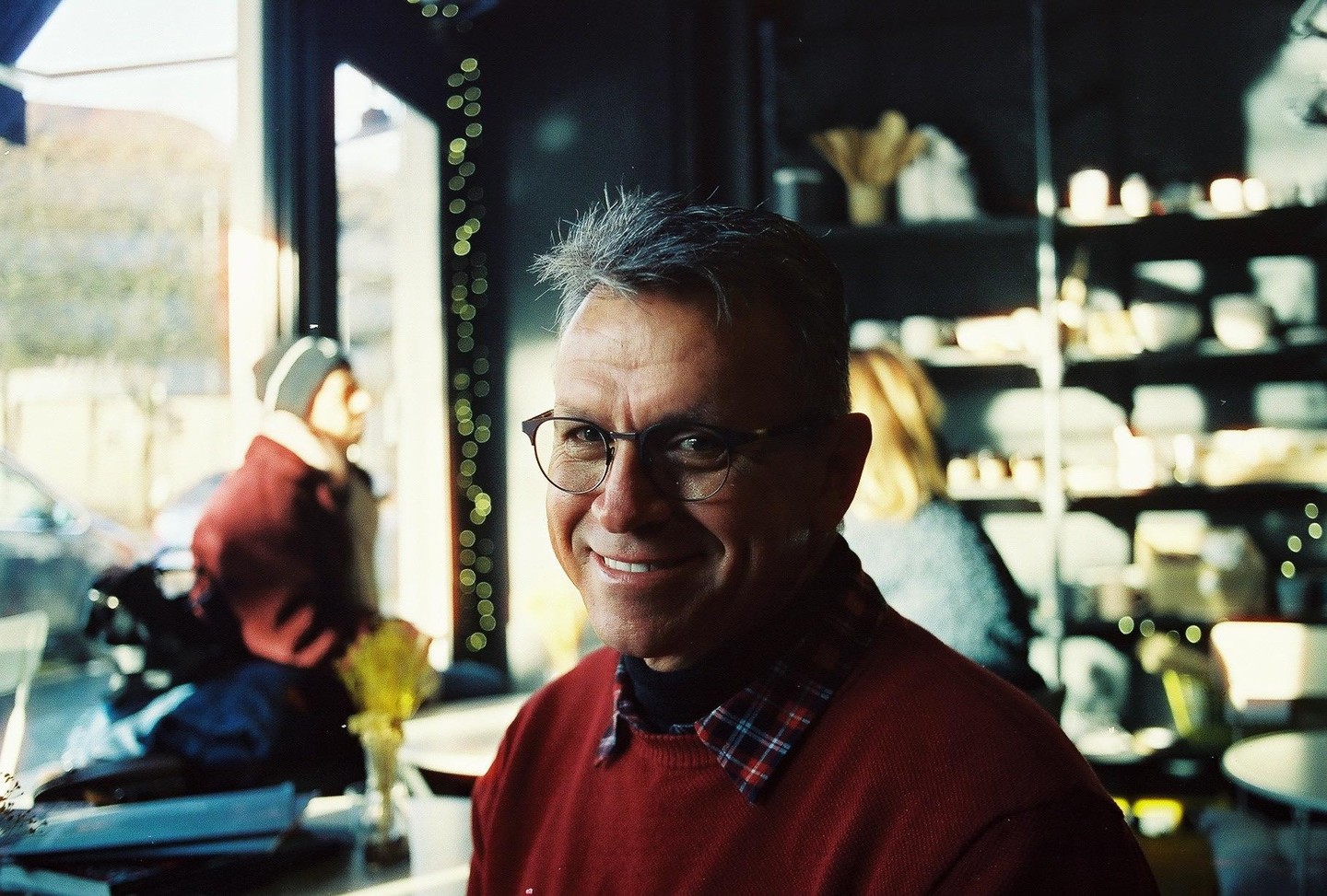 A man with glasses and a red sweater smiles at the camera in a cafe.