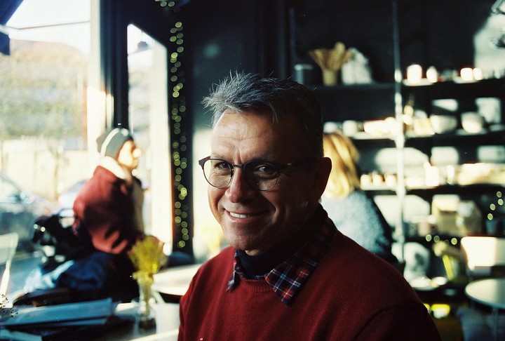 A man with glasses and a red sweater smiles at the camera in a cafe.
