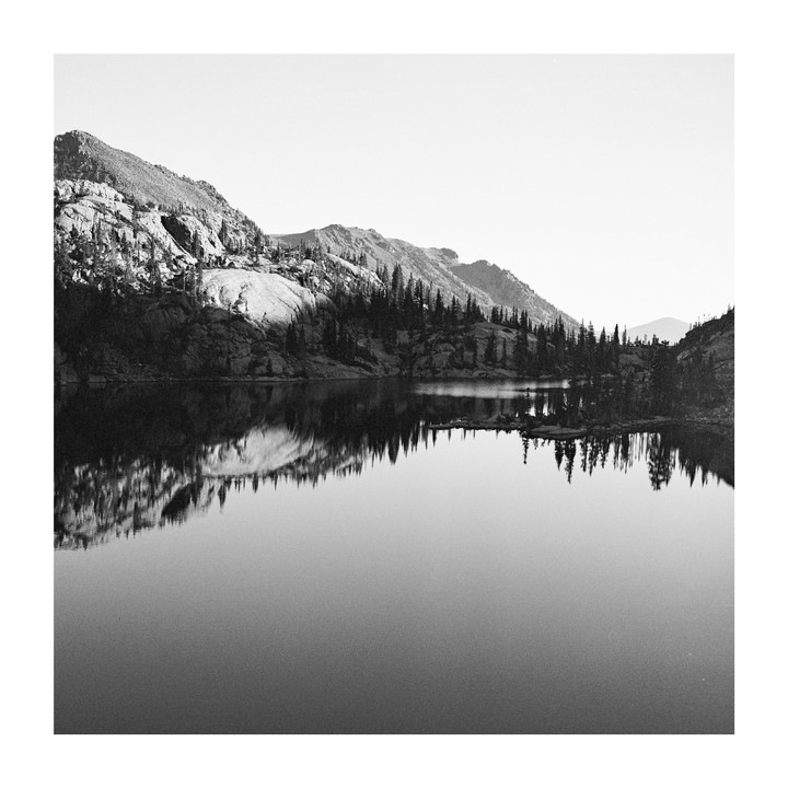 A still lake reflects mountains and a forest of pine trees under a bright sky.