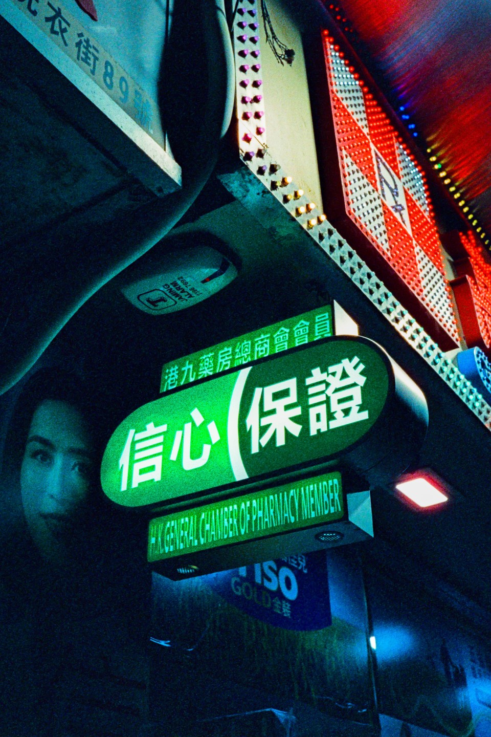 A green neon sign with Chinese characters and English text hangs above a street at night, with a partial reflection of a face visible on the left.