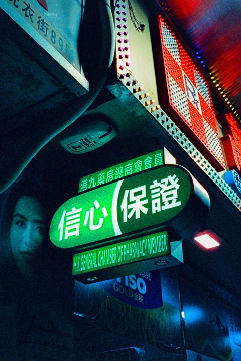 A green neon sign with Chinese characters and English text hangs above a street at night, with a partial reflection of a face visible on the left.