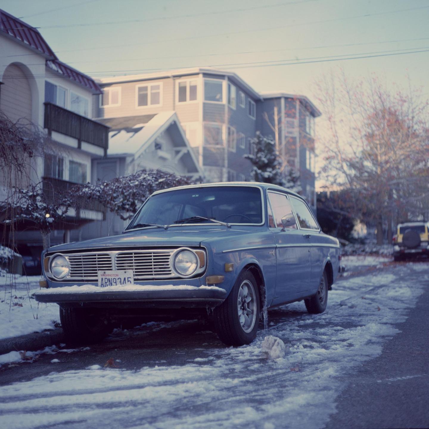 A blue car is parked on a snowy street in front of houses.