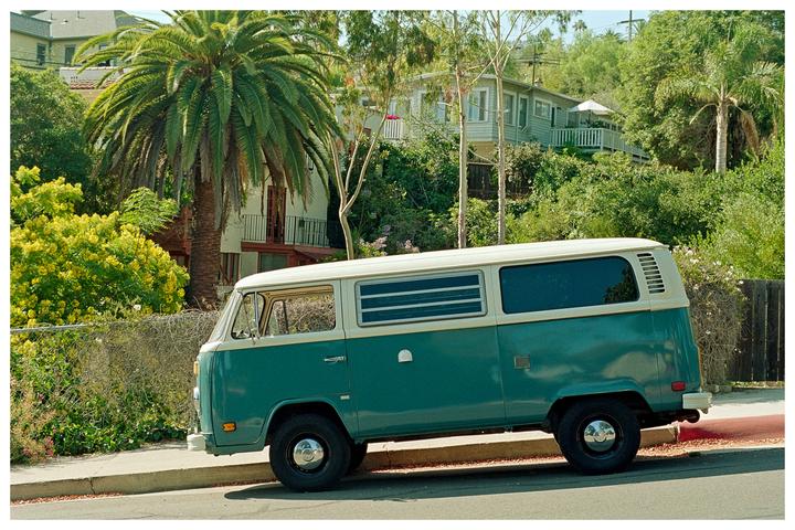 A teal and white van is parked on the side of a street lined with palm trees and houses.
