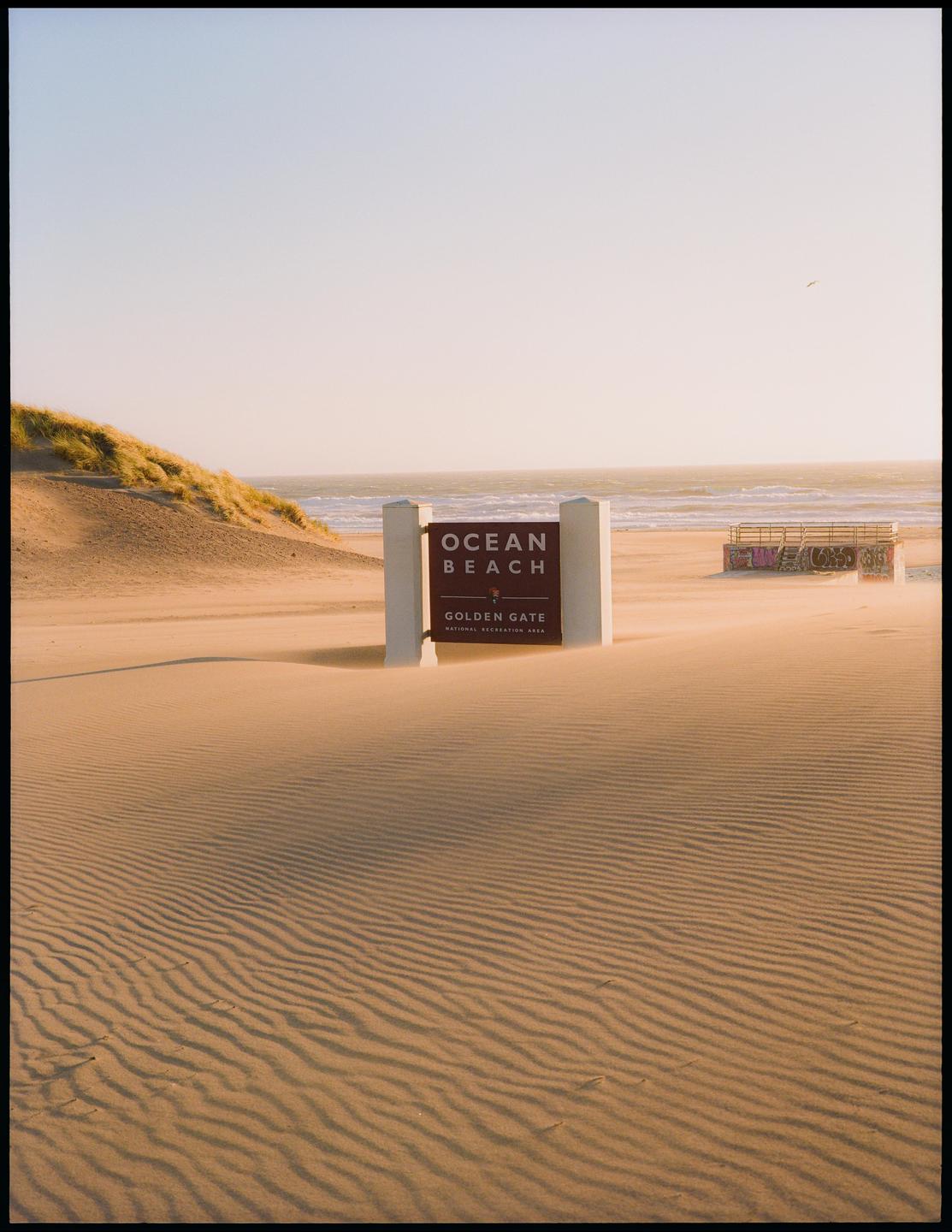 A sign for Ocean Beach and Golden Gate stands on a sandy beach with dunes and the ocean in the background.