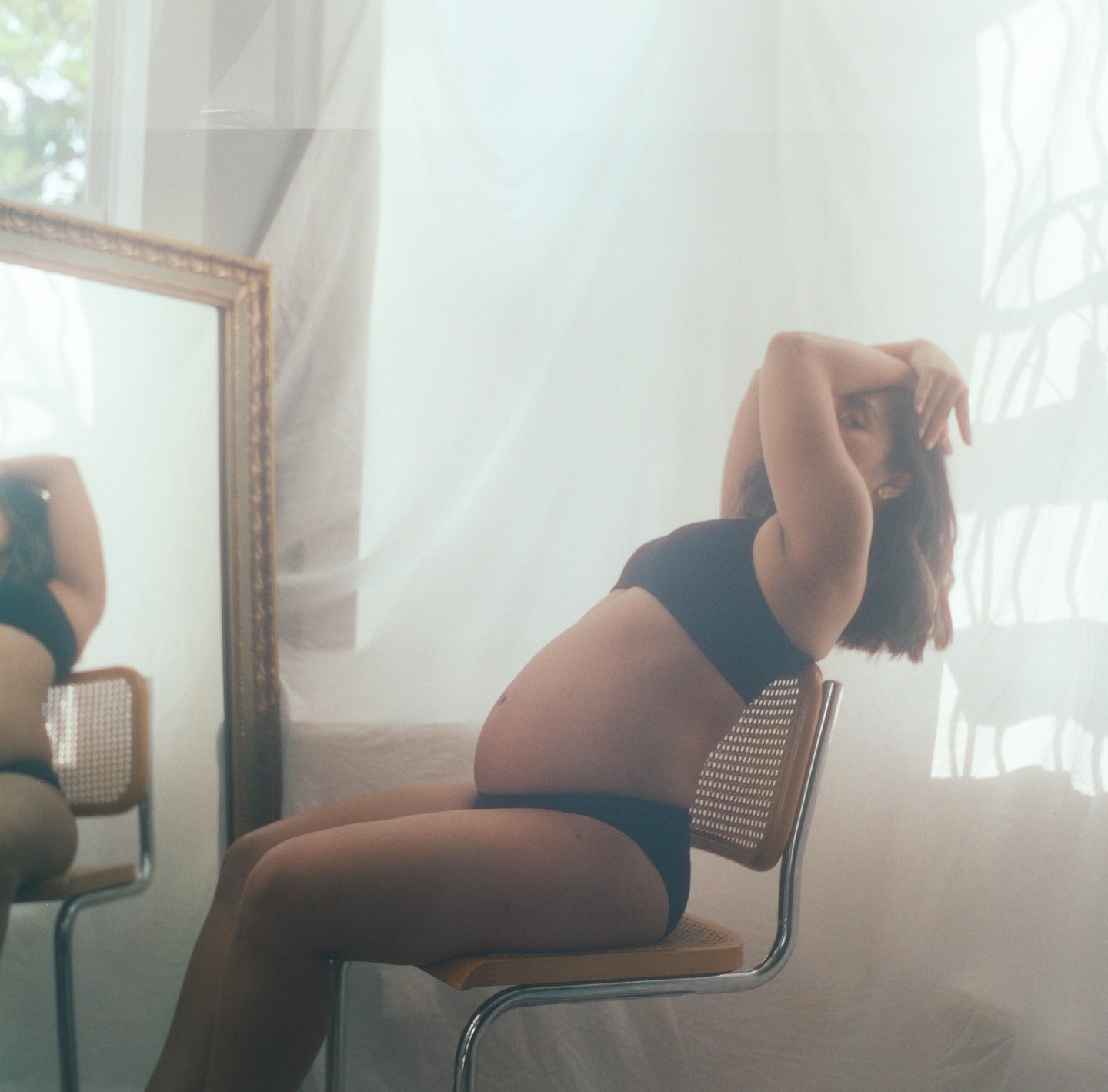 A pregnant woman sits on a chair with her arms above her head, looking at her reflection in a mirror.