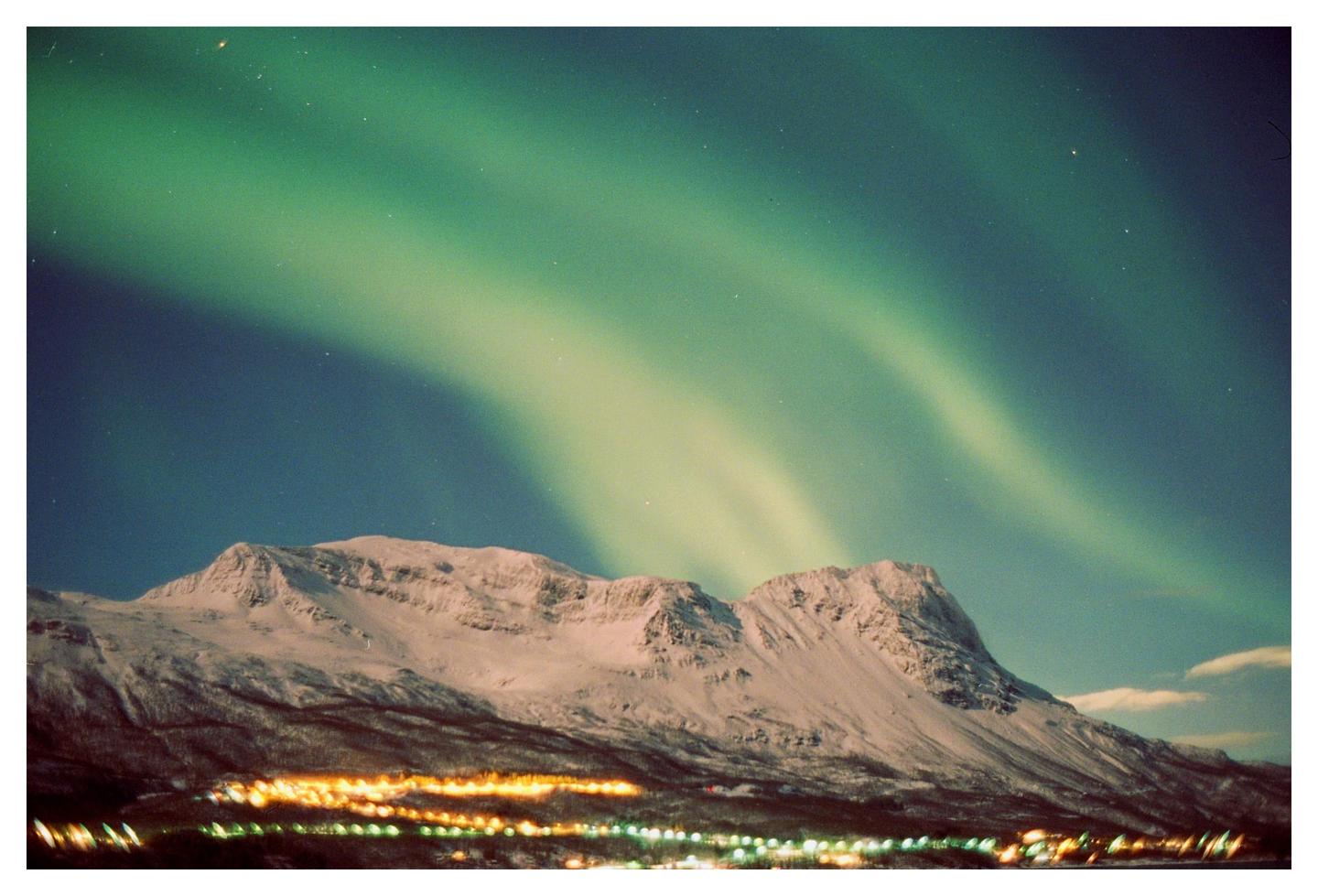 The aurora borealis glows green above a snow-covered mountain and a town at night.