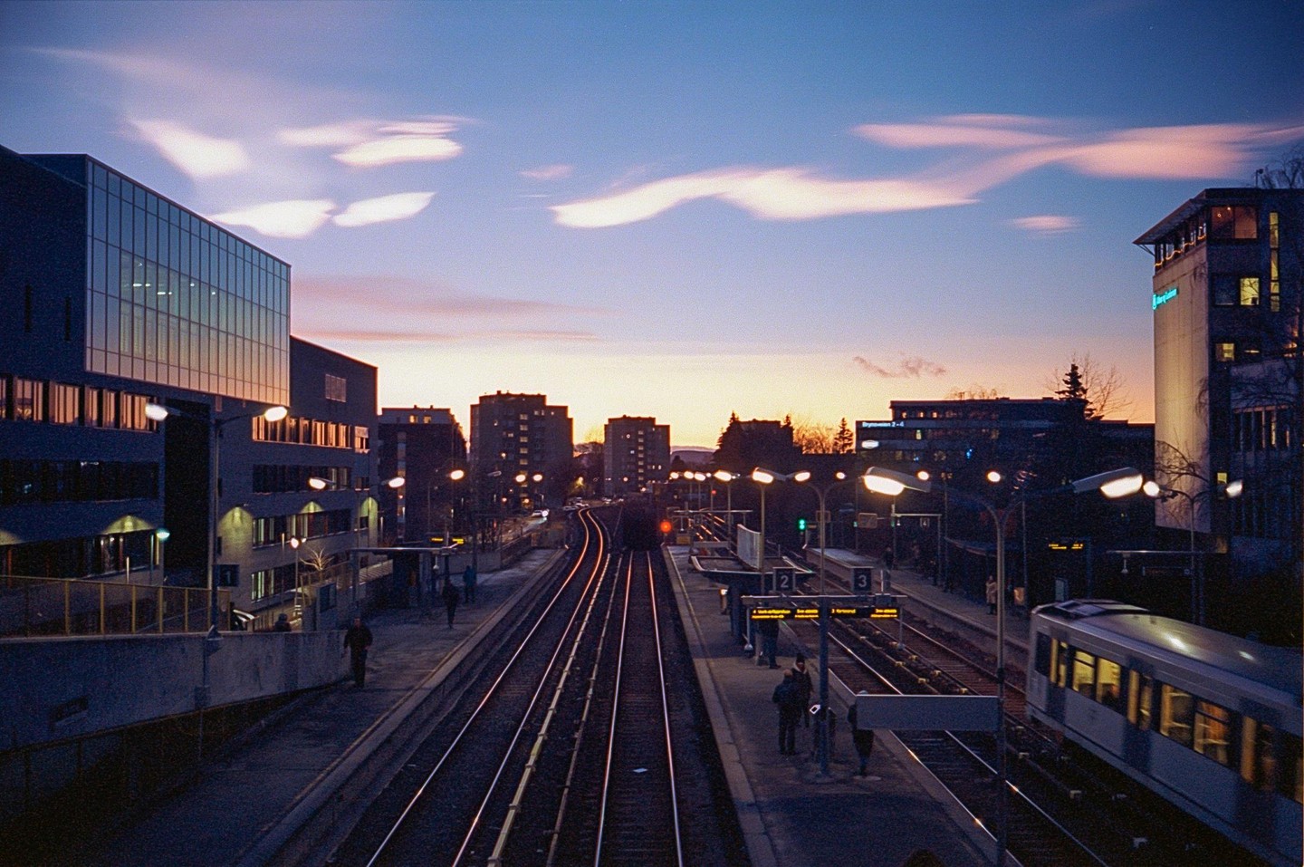 A train station with tracks and platforms is visible at dusk, with buildings on either side and a colorful sky overhead.