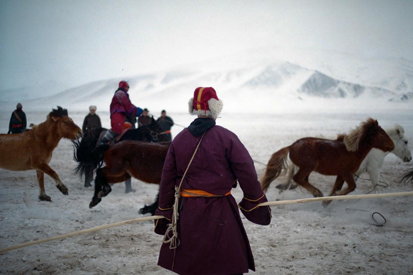 A person in traditional clothing stands in a snowy landscape with horses and other people in the background.