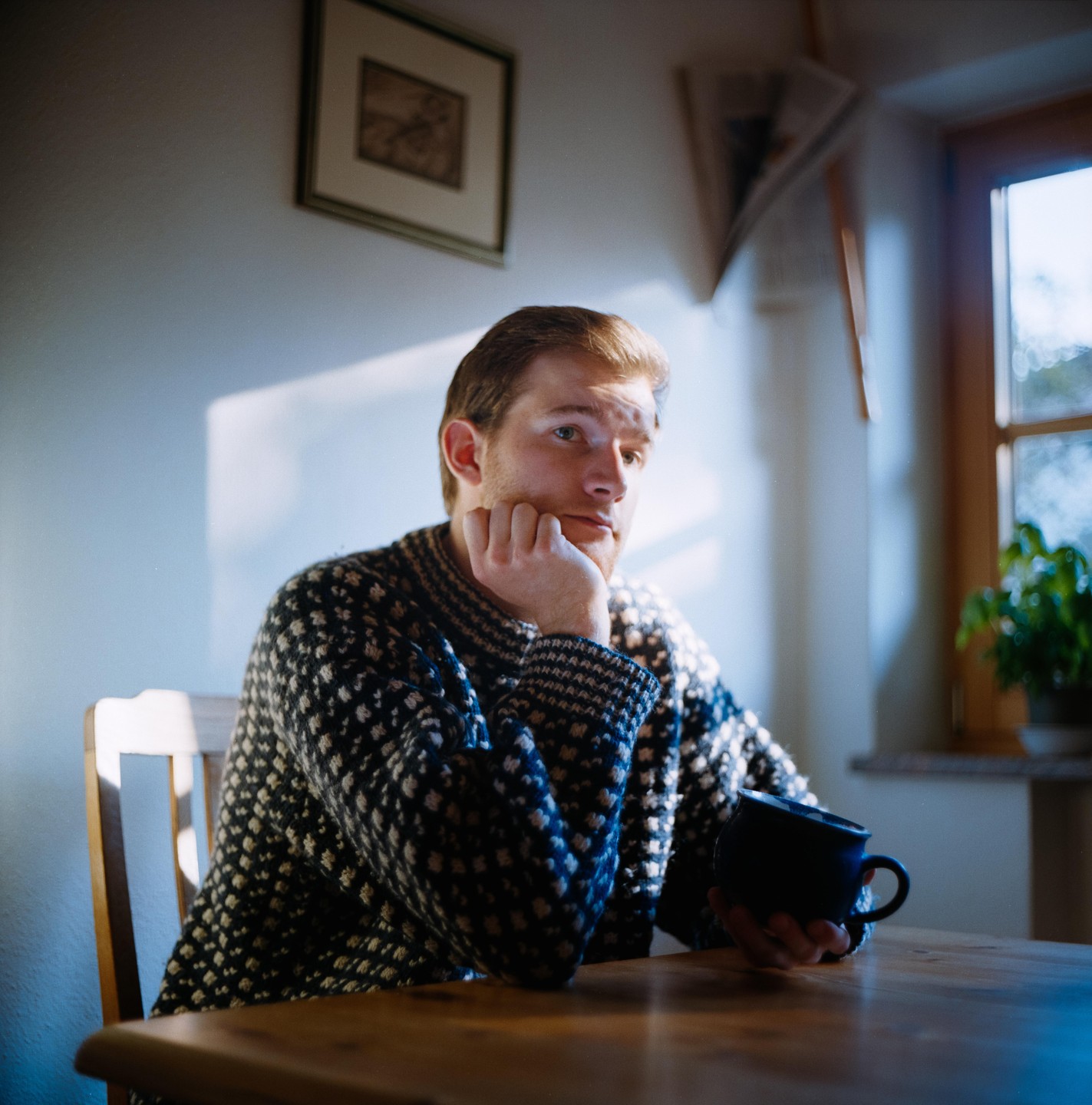 A man in a patterned sweater sits at a wooden table, holding a mug and looking out of frame.