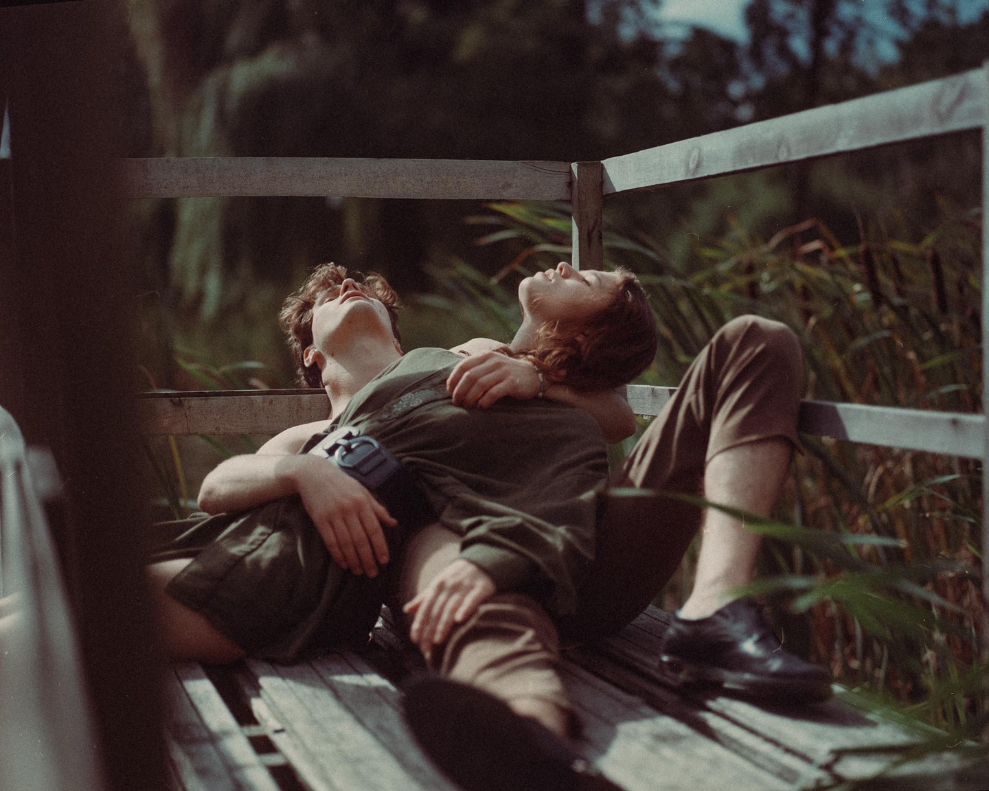 A couple is relaxing on a wooden bench with tall grass in the background.