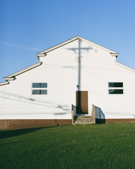A white building with a brown door and wooden steps has the shadow of a utility pole cast upon it.