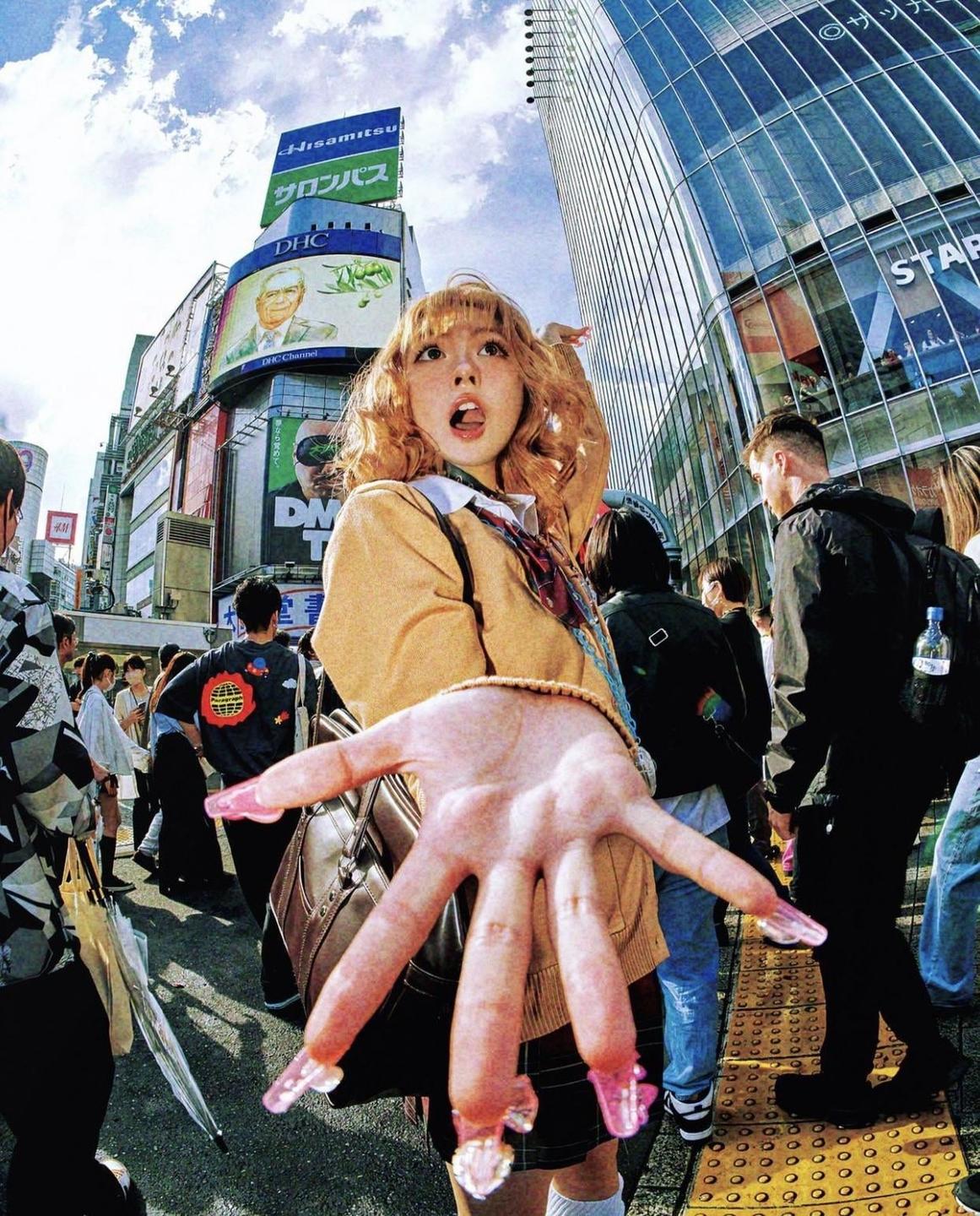 A woman with her tongue out and hand extended towards the viewer stands on a busy street with tall buildings and billboards.