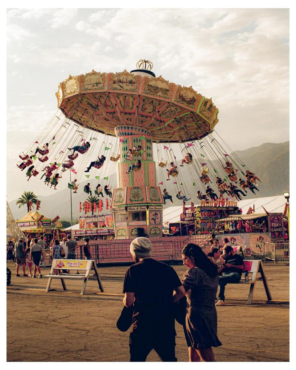 A swing ride with many people on it is visible at a fairground, with other fair attractions and people in the background.