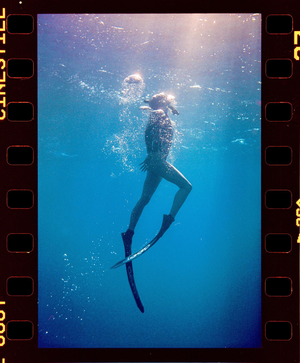 A woman wearing a snorkel mask and fins swims underwater in blue water with bubbles.
