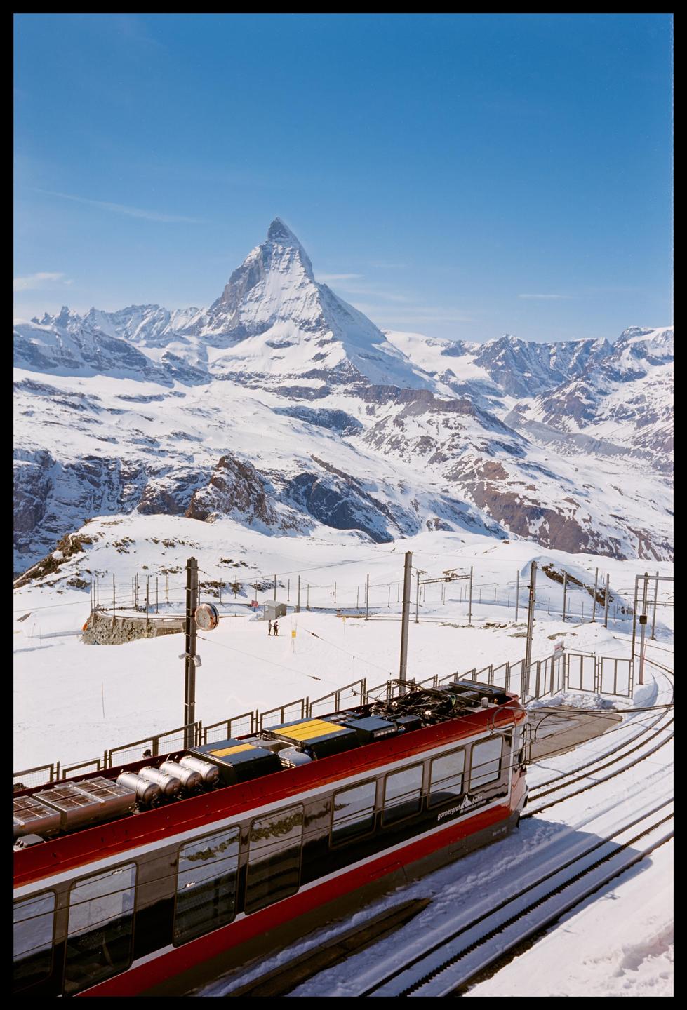 A red train is on tracks in a snowy mountain landscape with a prominent peak in the background.