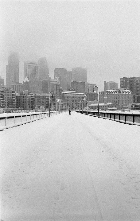A person walks on a snow-covered bridge towards a city skyline shrouded in fog.