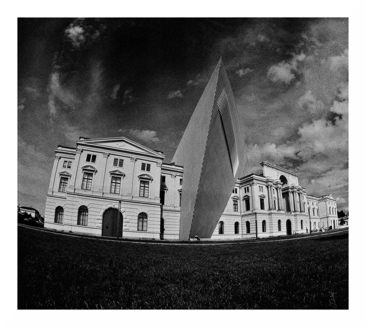 A modern building with a pointed roof stands between two classical buildings under a cloudy sky.
