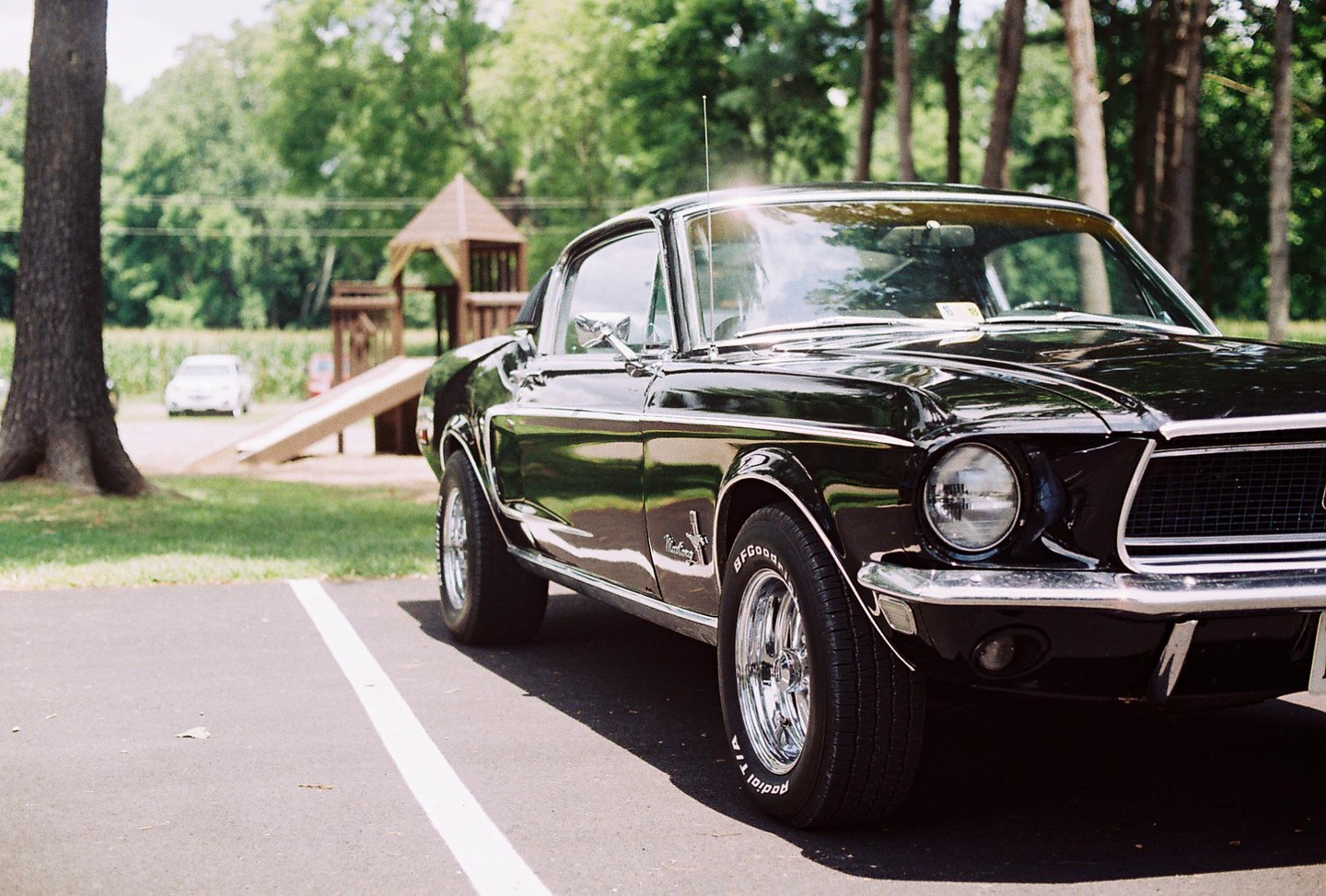 A black vintage car is parked on asphalt with a playground and trees in the background.