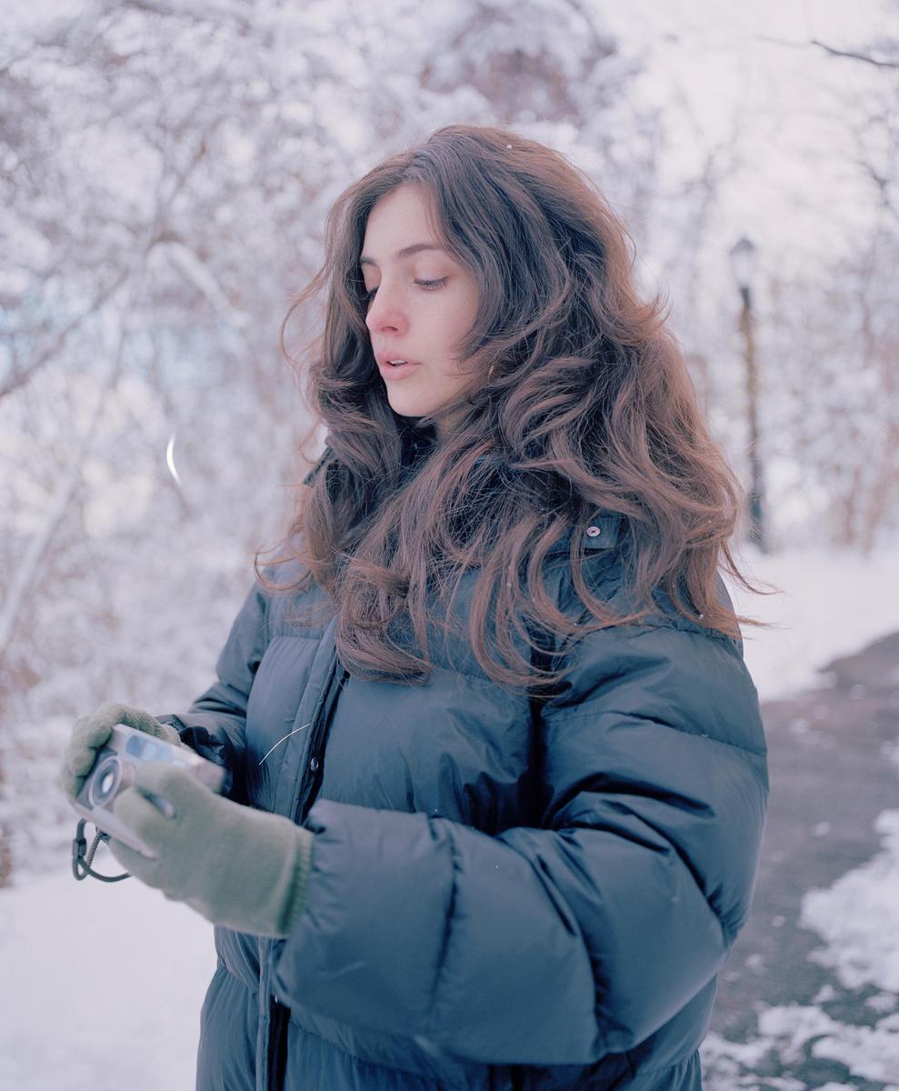 A woman wearing a puffer jacket and gloves holds a camera in a snowy landscape.