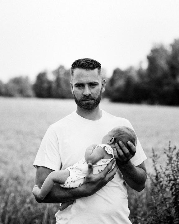 A man holds a baby in a field with trees in the background.