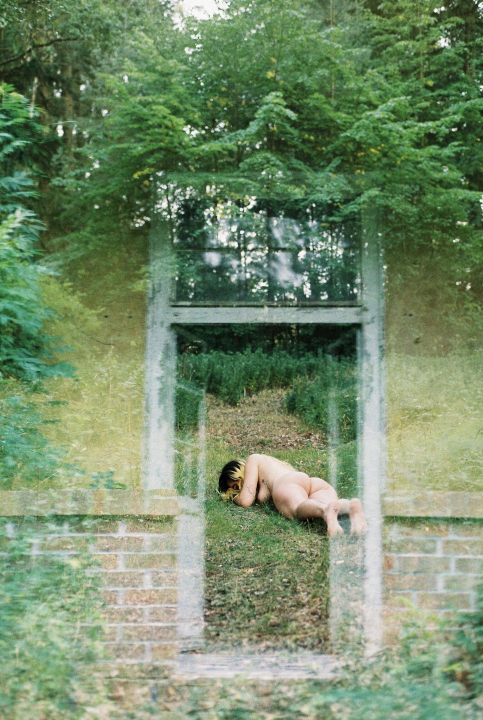 A nude woman lies on the grass in front of a brick wall with a window and a door frame, with trees visible through a double exposure.