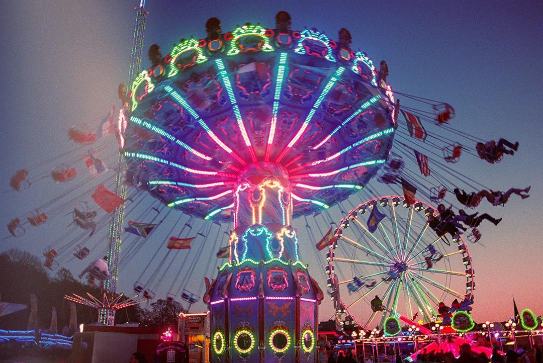 A brightly lit swing ride and ferris wheel are visible against a dusk sky at a fairground.