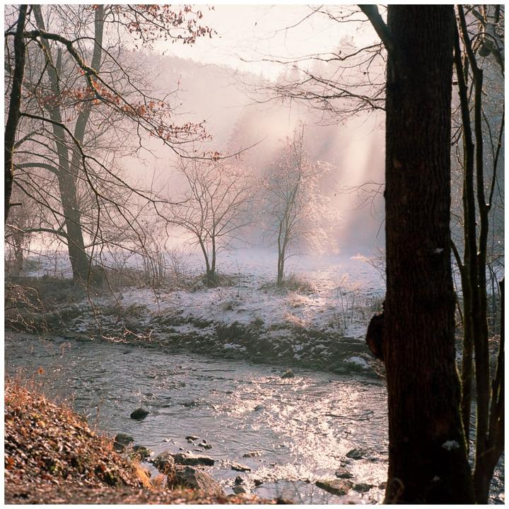 A river flows through a snowy landscape with trees and hazy light in the background.