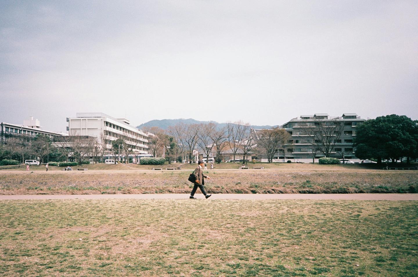 A man walks across a grassy field with buildings and trees in the background.