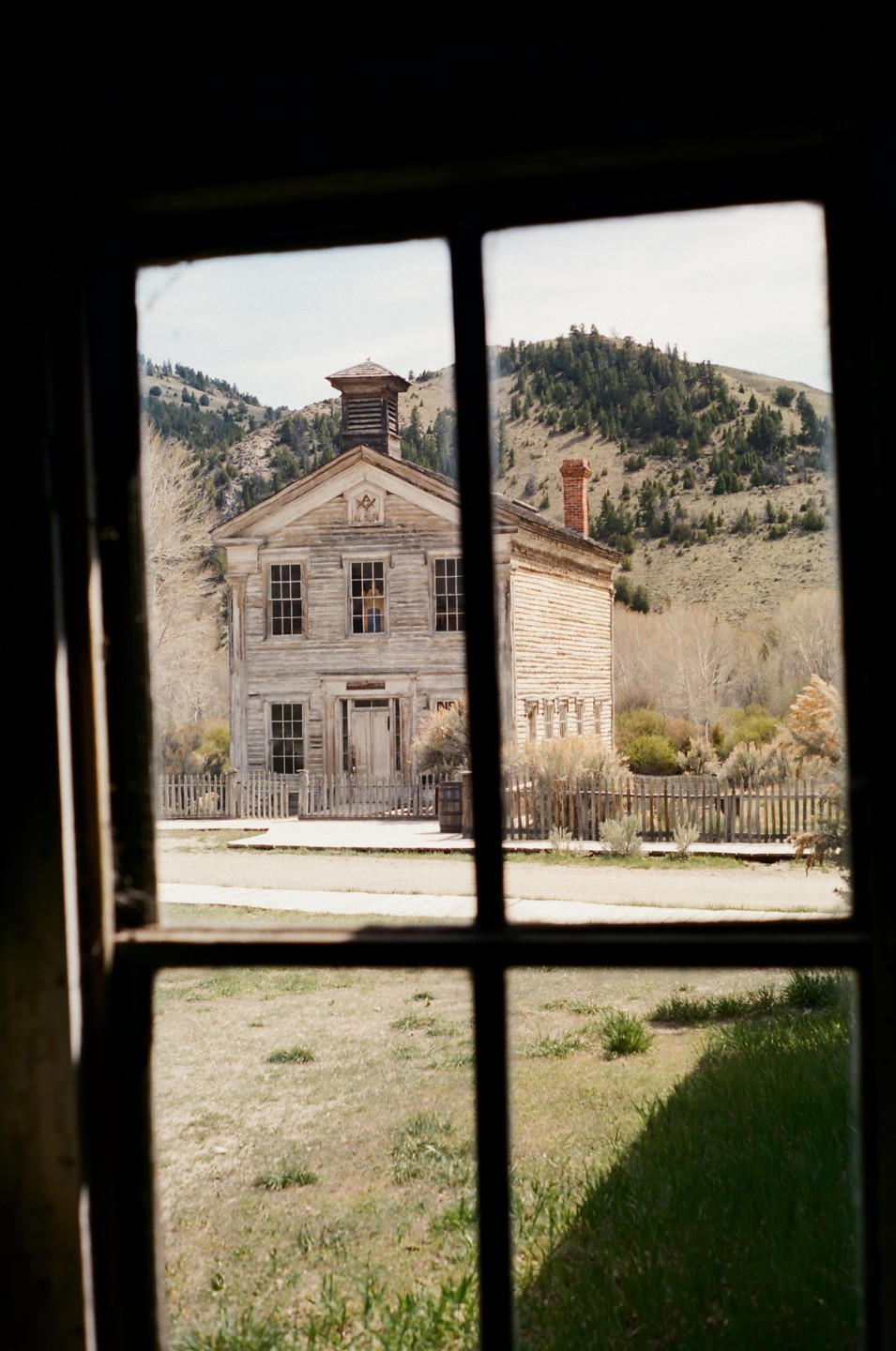 A weathered wooden building with a cupola and a red brick chimney stands in a grassy field, viewed through a window.