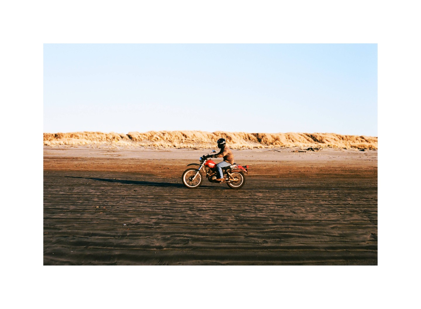 A person rides a red motorcycle on a sandy beach with dunes in the background.