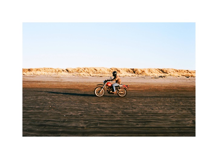 A person rides a red motorcycle on a sandy beach with dunes in the background.