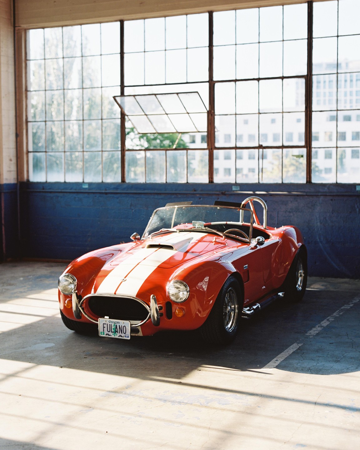 A red Shelby Cobra replica with white racing stripes is parked in a garage with large windows.