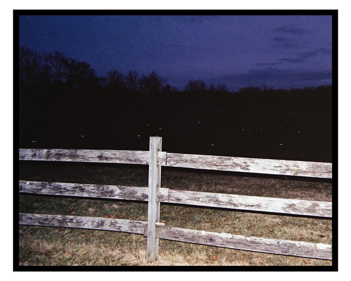 A wooden fence stands in front of a dark field with glowing animal eyes and a dark blue sky.