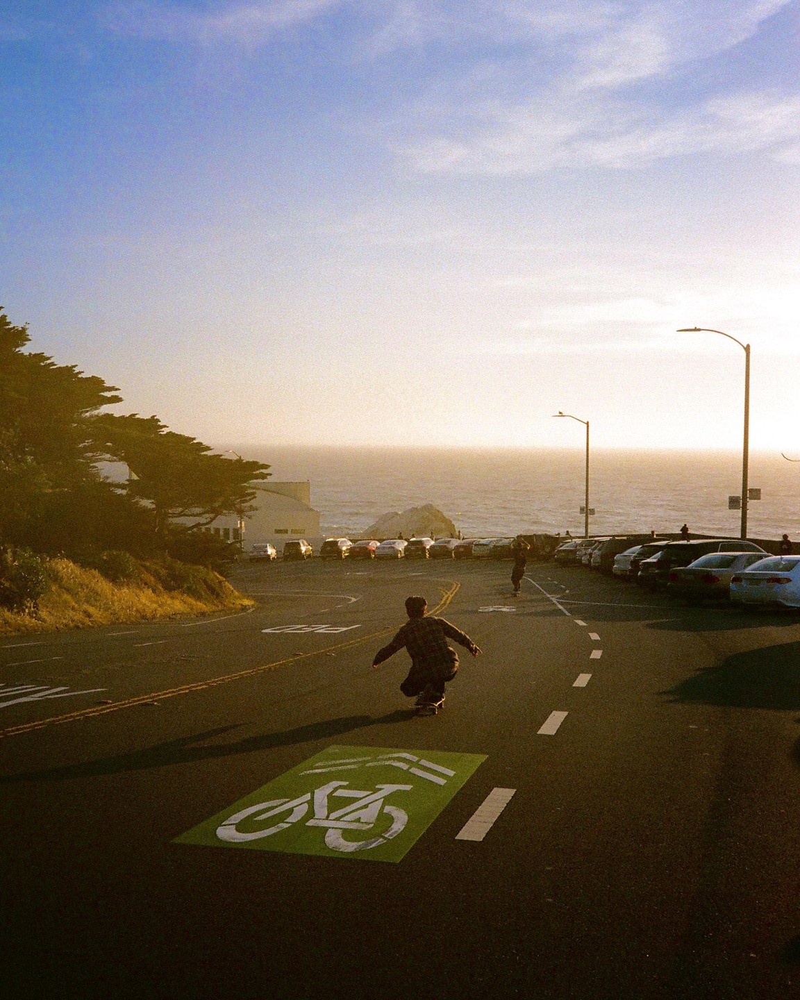 A person on a skateboard rides down a road with a bicycle lane symbol painted on it, towards the ocean at sunset.