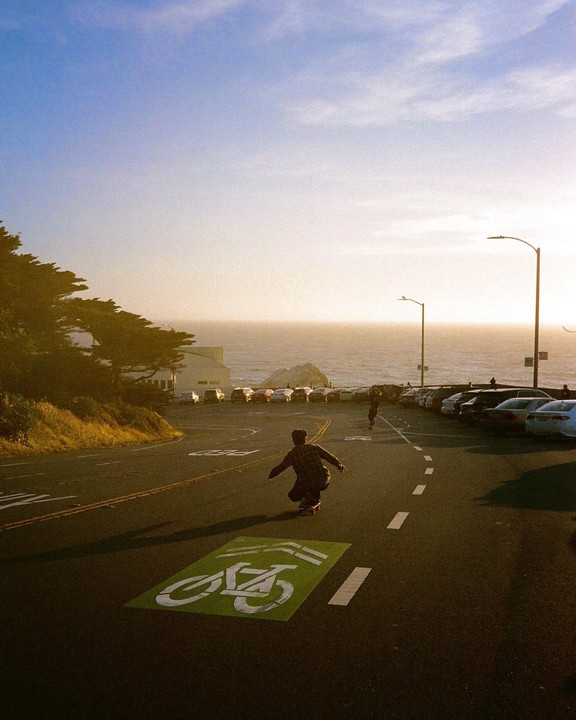 A person on a skateboard rides down a road with a bicycle lane symbol painted on it, towards the ocean at sunset.