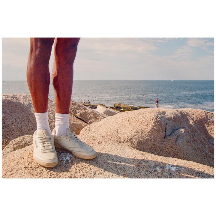 A person stands on a rocky shore overlooking the ocean with a sailboat in the distance.