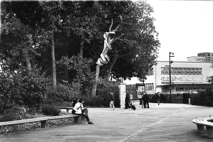 A black and white street scene shows people walking and sitting on a bench, with a tree wrapped in fabric in the background.