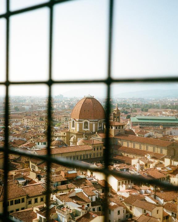 A grid of bars frames a cityscape with a large domed building and many terracotta roofs under a hazy sky.