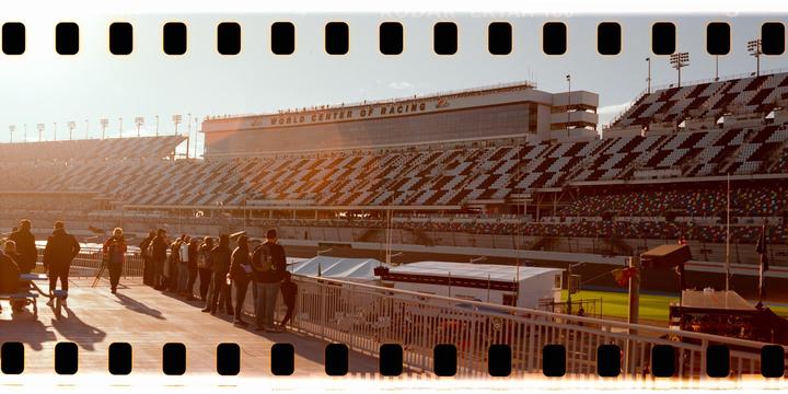 A crowd of people stands on a walkway overlooking a large stadium with empty seats and a building labeled "World Center of Racing" in the background.