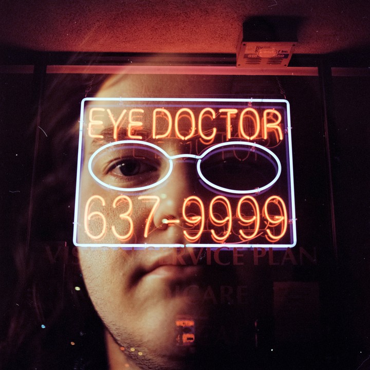A man's face is visible behind a neon sign for an eye doctor, with the sign's glasses shape aligned over his eye.