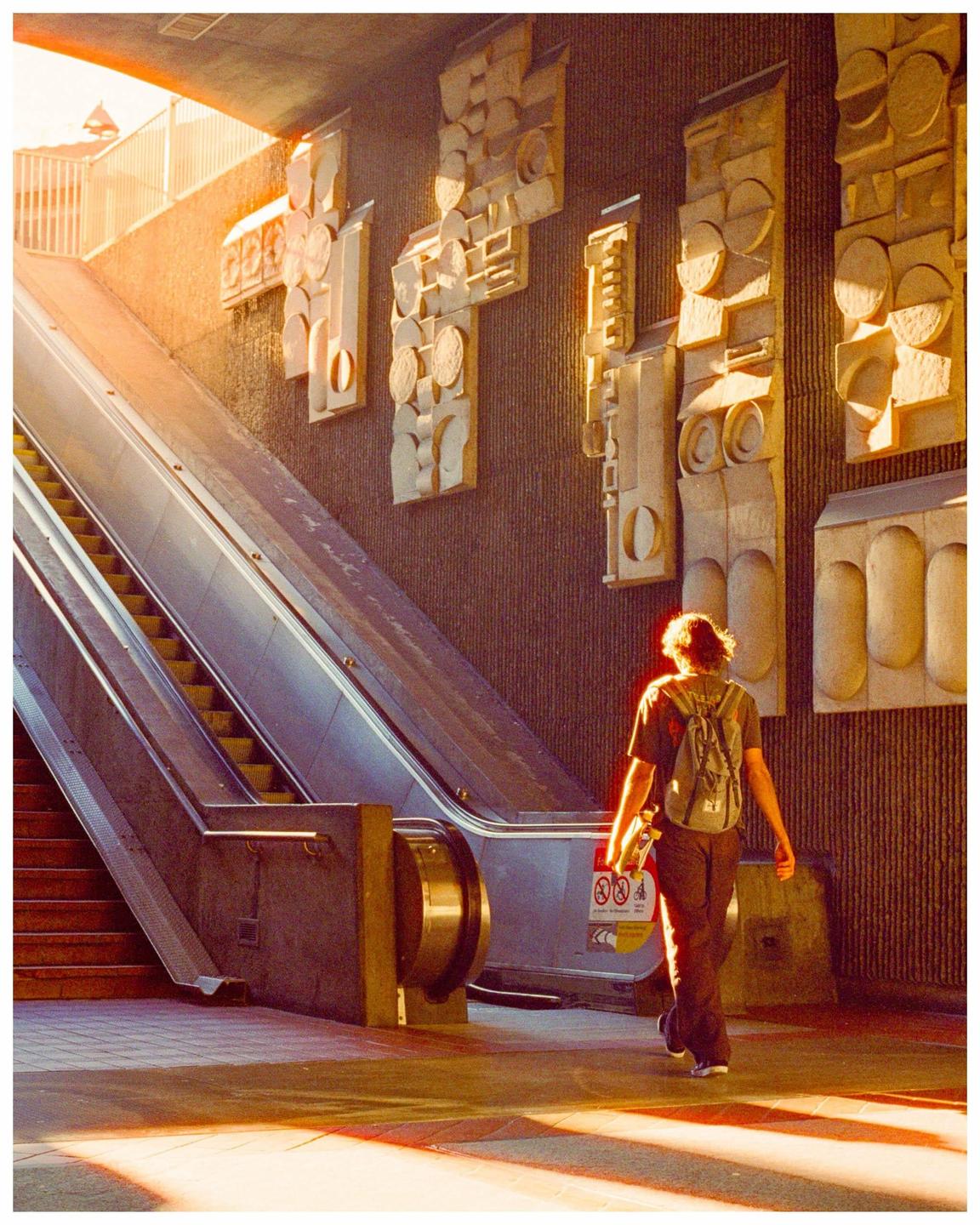 A man with a backpack and skateboard walks away from an escalator in a BART station.