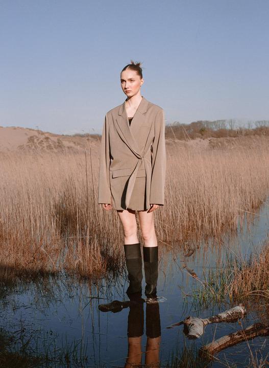 A woman in a blazer and tall boots stands in shallow water surrounded by tall dry grass.