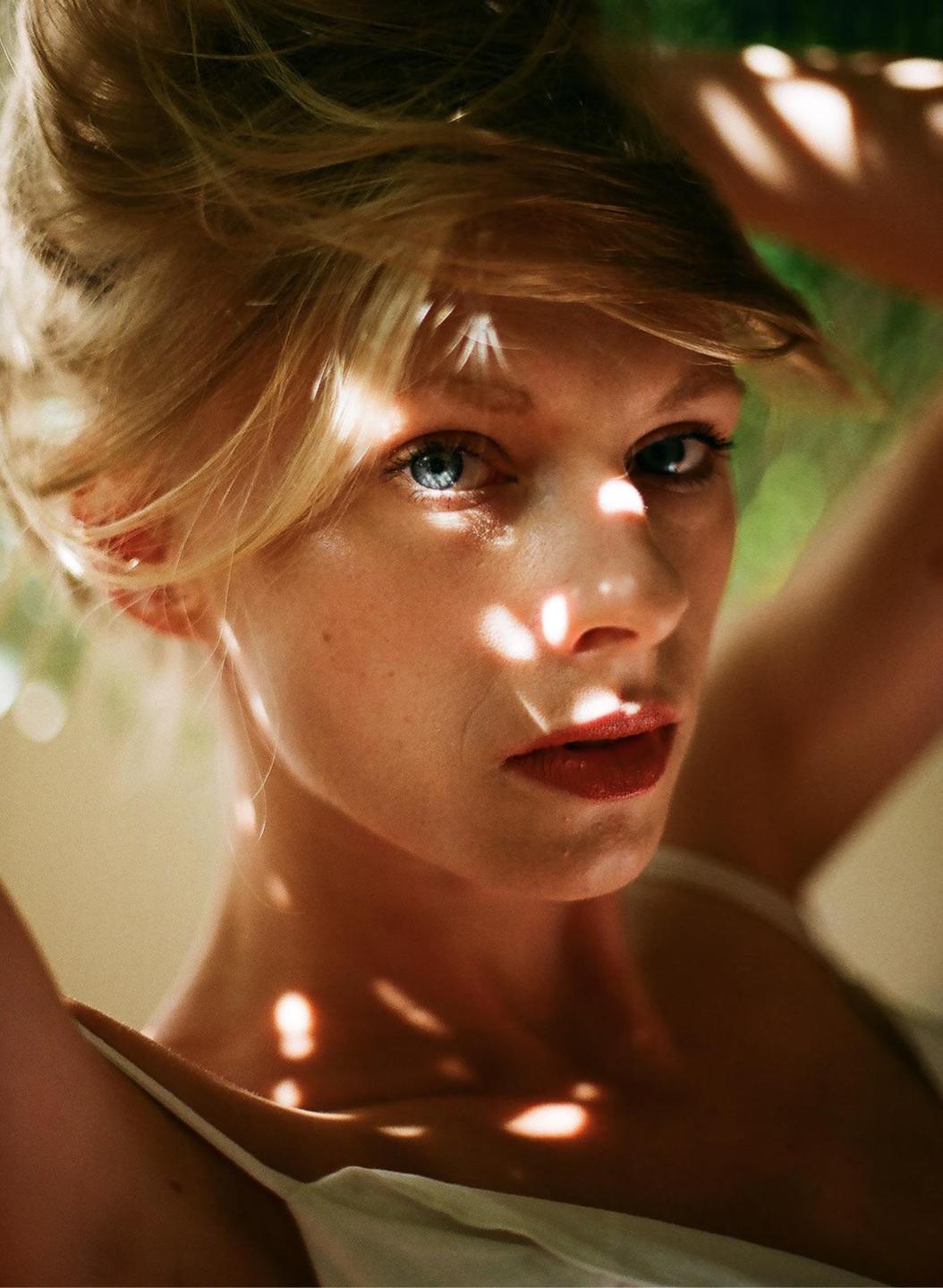 A woman with blonde hair and blue eyes looks directly at the viewer, with dappled sunlight on her face and shoulders.