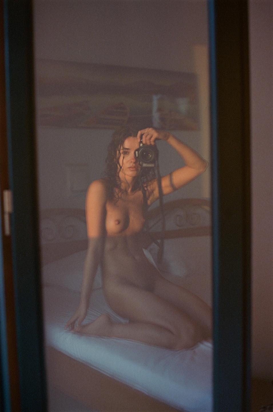 A woman kneels on a bed, reflected in a mirror, holding a camera to take a self-portrait.